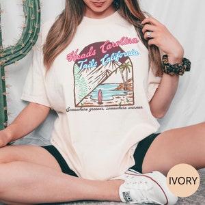 May include: A white t-shirt with a graphic design of a beach scene and the text "Heads Carolina Tails California" in pink, blue, and brown. The text "Somewhere greener, somewhere warmer" is printed below the graphic. The shirt is labeled "IVORY".