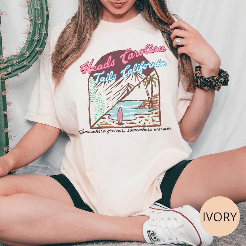 May include: A white t-shirt with a graphic design of a beach scene and the text "Heads Carolina Tails California" in pink, blue, and brown. The text "Somewhere greener, somewhere warmer" is printed below the graphic. The shirt is labeled "IVORY".