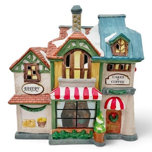 May include: A ceramic miniature bakery building with a snow-dusted roof, featuring a red and white striped awning, green trim, and signs that read BAKERY and CAKES & COFFEE. The structure includes multiple windows, a chimney, and a small door.