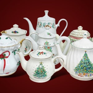Op de afbeelding: Een verzameling witte keramische theepotten met diverse kerstthema-ontwerpen, waaronder versierde kerstbomen, rode linten, hulstbladeren en bessen. Elke theepot heeft een deksel, een handvat en een tuit, sommige met een gouden rand.