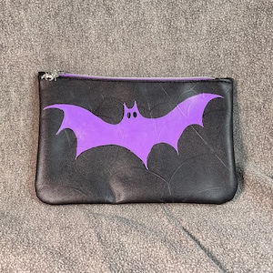 May include: A black zippered pouch with a purple bat design. The bat has two small white eyes.