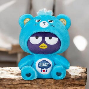 May include: A plush toy featuring a blue bear wearing a blue hooded outfit with a purple face and yellow beak. The bear has a sad expression and the name "Stacy" is on its chest. The toy is sitting on a wooden surface.