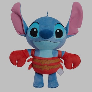 May include: A plush toy of the character Stitch from Lilo & Stitch. The blue alien has large ears, big black eyes, and a smiling expression. He wears a red crab-like outfit with brown rope detailing. The toy is standing upright.