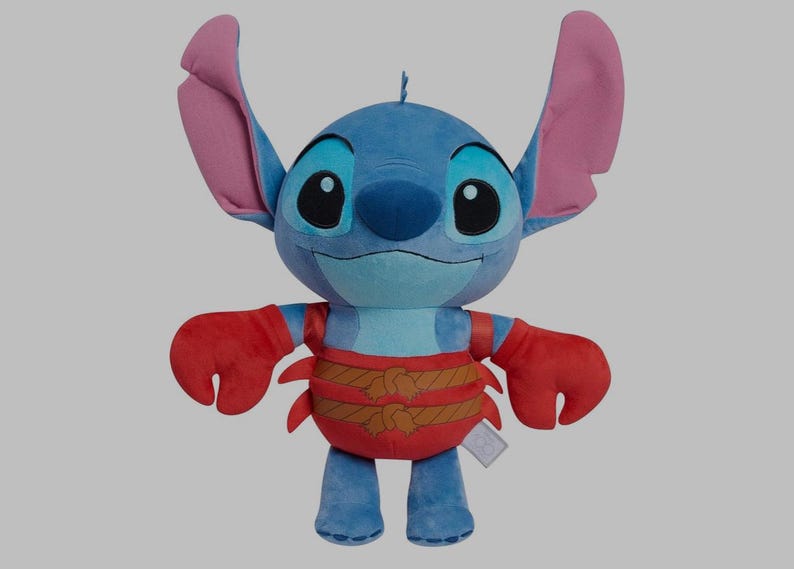 May include: A plush toy of the character Stitch from Lilo & Stitch. The blue alien has large ears, big black eyes, and a smiling expression. He wears a red crab-like outfit with brown rope detailing. The toy is standing upright.