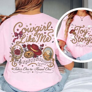 Pu&ograve; includere: Una t-shirt rosa chiaro con un design grafico sul retro con il testo Cowgirl Like Me, Eyes full of Stars e Takes One to Know One, insieme a illustrazioni di un lazo, cappello da cowboy, rose e una scena del deserto. Il davanti mostra Toy Story.