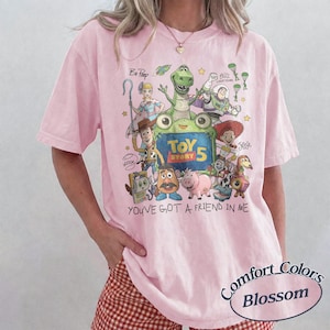 May include: A light pink t-shirt featuring a colorful graphic of Toy Story characters including Woody, Buzz Lightyear, Jessie, and Rex. The design includes the text Toy Story 5 and You've Got A Friend In Me. The shirt has short sleeves and a crew neckline.