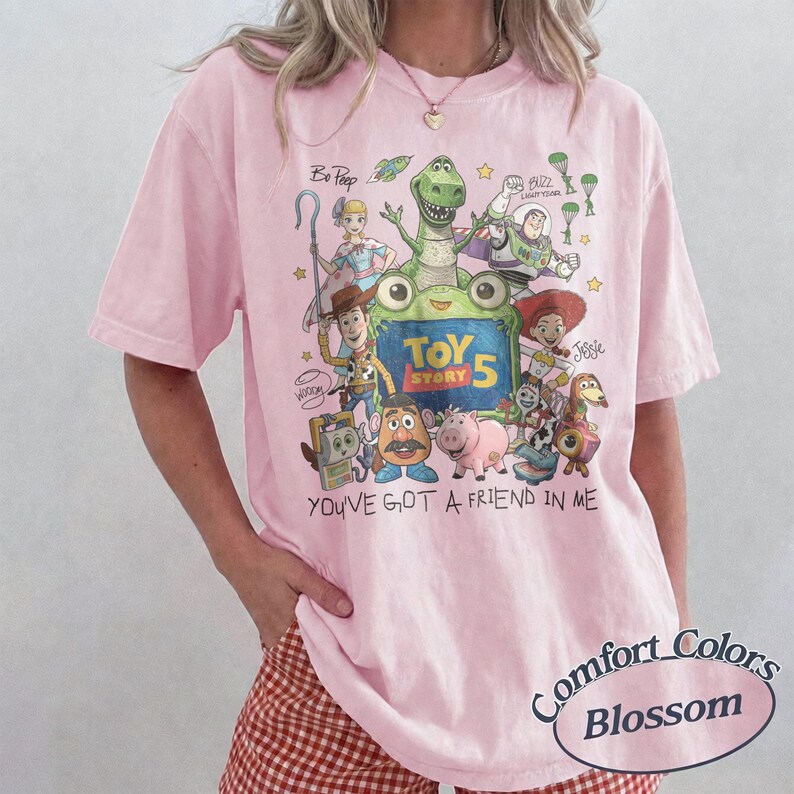 May include: A light pink t-shirt featuring a colorful graphic of Toy Story characters including Woody, Buzz Lightyear, Jessie, and Rex. The design includes the text Toy Story 5 and You've Got A Friend In Me. The shirt has short sleeves and a crew neckline.