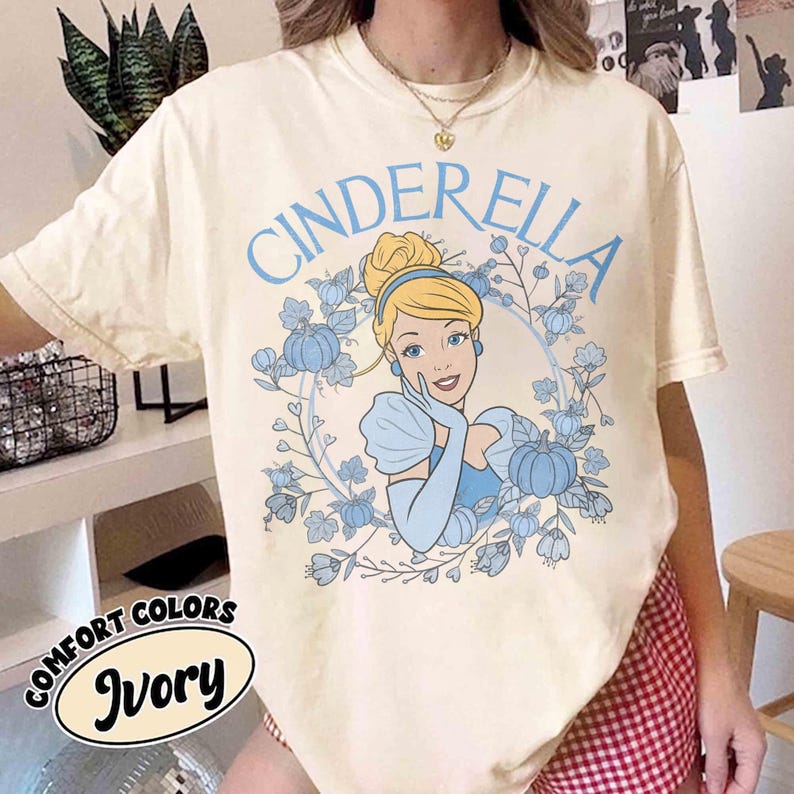 May include: An ivory short-sleeve t-shirt featuring a graphic print of Cinderella in a blue dress surrounded by blue pumpkins and floral vines. The word CINDERELLA is printed in blue arched text above the character. The shirt has a crew neckline.