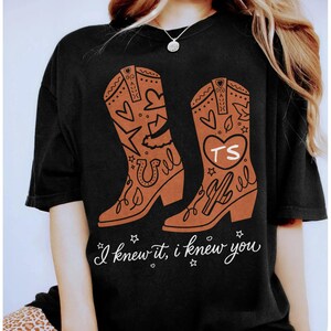 May include: A black short-sleeved t-shirt featuring a graphic of two brown cowboy boots decorated with hearts, stars, a horseshoe, and a cactus. One boot displays the letters TS inside a heart. The text I knew it, i knew you is printed below.