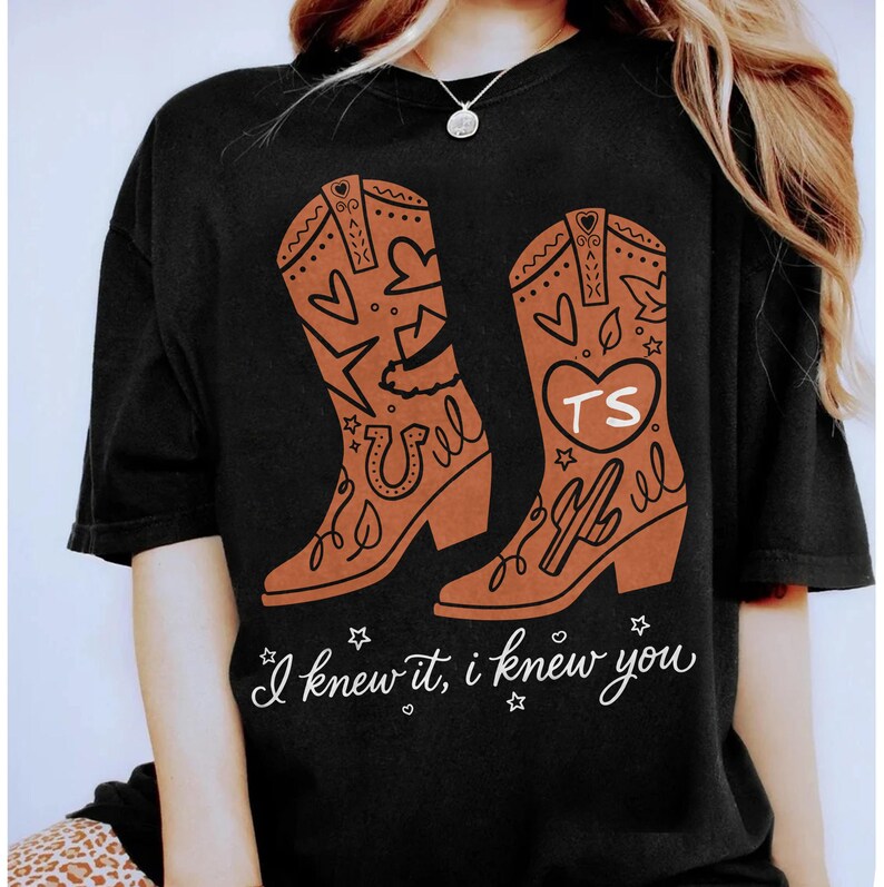 May include: A black short-sleeved t-shirt featuring a graphic of two brown cowboy boots decorated with hearts, stars, a horseshoe, and a cactus. One boot displays the letters TS inside a heart. The text I knew it, i knew you is printed below.