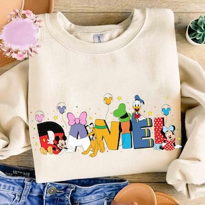 Könnte beinhalten: Cremefarbenes Sweatshirt mit dem Namen "DANIEL" in bunten Blockbuchstaben. Micky Maus, Pluto, Donald Duck und Minnie Maus sind mit Luftballons abgebildet. Das Sweatshirt liegt auf einer Holzoberfläche mit Jeans.