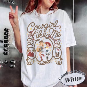 May include: A white t-shirt featuring a brown rope graphic framing the text Cowgirl like Me. Inside the rope are illustrations of a woman playing guitar, a cowgirl character, a red jacket, a cowboy hat, and a cowboy boot with a microphone.