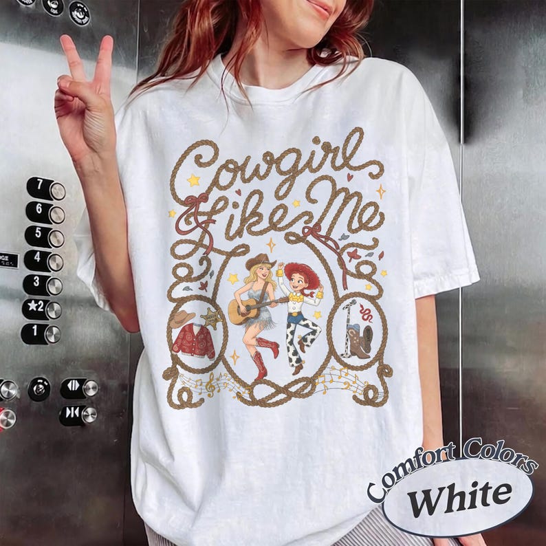 May include: A white t-shirt featuring a brown rope graphic framing the text Cowgirl like Me. Inside the rope are illustrations of a woman playing guitar, a cowgirl character, a red jacket, a cowboy hat, and a cowboy boot with a microphone.
