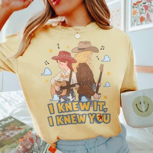 May include: A pale yellow t-shirt featuring a graphic of two figures in cowboy hats playing guitars. The design includes musical notes, stars, and clouds, with the text "I KNEW IT, I KNEW YOU" printed in bold blue letters at the bottom of the illustration.