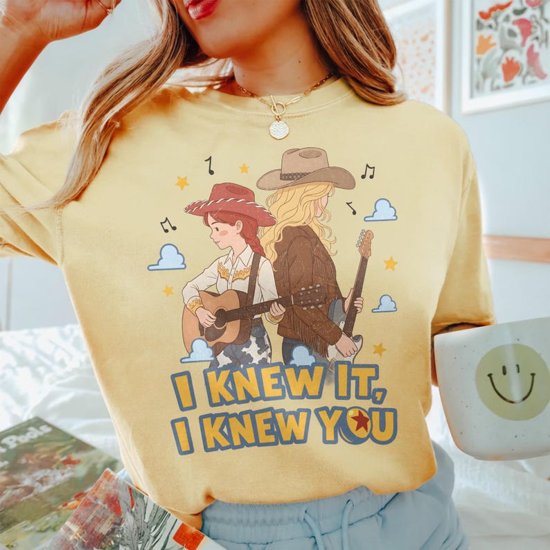 May include: A pale yellow t-shirt featuring a graphic of two figures in cowboy hats playing guitars. The design includes musical notes, stars, and clouds, with the text "I KNEW IT, I KNEW YOU" printed in bold blue letters at the bottom of the illustration.