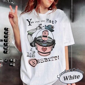 Puede incluir: Camiseta blanca con un gr&aacute;fico de dibujos animados de un personaje con un traje espacial rosa y verde, un sombrero azul y el texto "You See the Hat? I Am Mrs. Nesbitt!" La camiseta tambi&eacute;n tiene las palabras "Comfort Colors White".