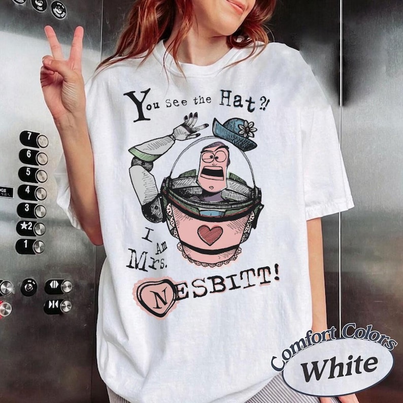Puede incluir: Camiseta blanca con un gr&aacute;fico de dibujos animados de un personaje con un traje espacial rosa y verde, un sombrero azul y el texto "You See the Hat? I Am Mrs. Nesbitt!" La camiseta tambi&eacute;n tiene las palabras "Comfort Colors White".