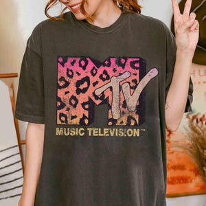 May include: Dark gray t-shirt featuring a retro MTV logo with a pink and orange leopard print background. The words "MUSIC TELEVISION" are printed below the logo in a gold color. The shirt has a vintage, worn look.