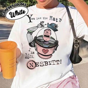 Puede incluir: Camiseta blanca con un dibujo animado de un personaje en un cubo rosa con el texto "You see the Hat? I am Mrs. NESBITT!". La camiseta tambi&eacute;n tiene las palabras "COMFORT COLORS" y "White" en un &oacute;valo blanco.