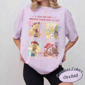 May include: A light purple t-shirt featuring four rectangular panels with cartoon illustrations of characters and a cowboy boot. The text reads, Love has way BRINGING THINGS BACK TO LIFE. The design includes pink bows, hearts, and colorful blocks.