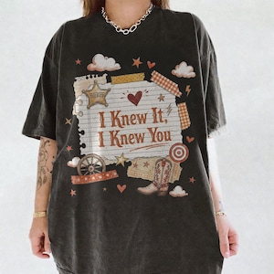 May include: A black short-sleeve t-shirt featuring a graphic print of a lined notepad page with the text "I Knew It, I Knew You" in brown. The design includes a sheriff star, a cowboy boot, a wagon wheel, clouds, hearts, and stars in warm tones.