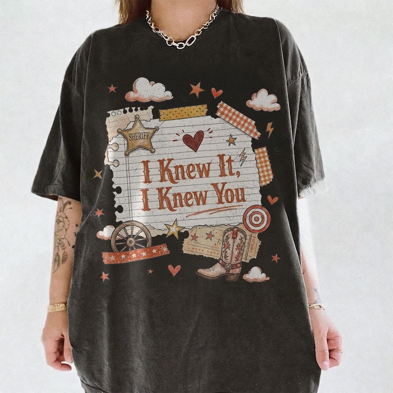 May include: A black short-sleeve t-shirt featuring a graphic print of a lined notepad page with the text "I Knew It, I Knew You" in brown. The design includes a sheriff star, a cowboy boot, a wagon wheel, clouds, hearts, and stars in warm tones.