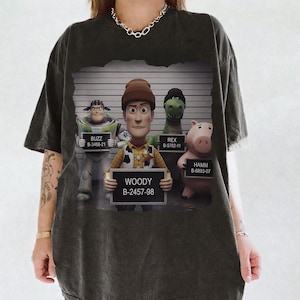 Puede incluir: Una camiseta gris carb&oacute;n con un estampado gr&aacute;fico de cuatro personajes de Toy Story posando para una foto policial frente a una tabla de altura. Los personajes Buzz, Woody, Rex y Hamm sostienen carteles con sus nombres y n&uacute;meros de identificaci&oacute;n.