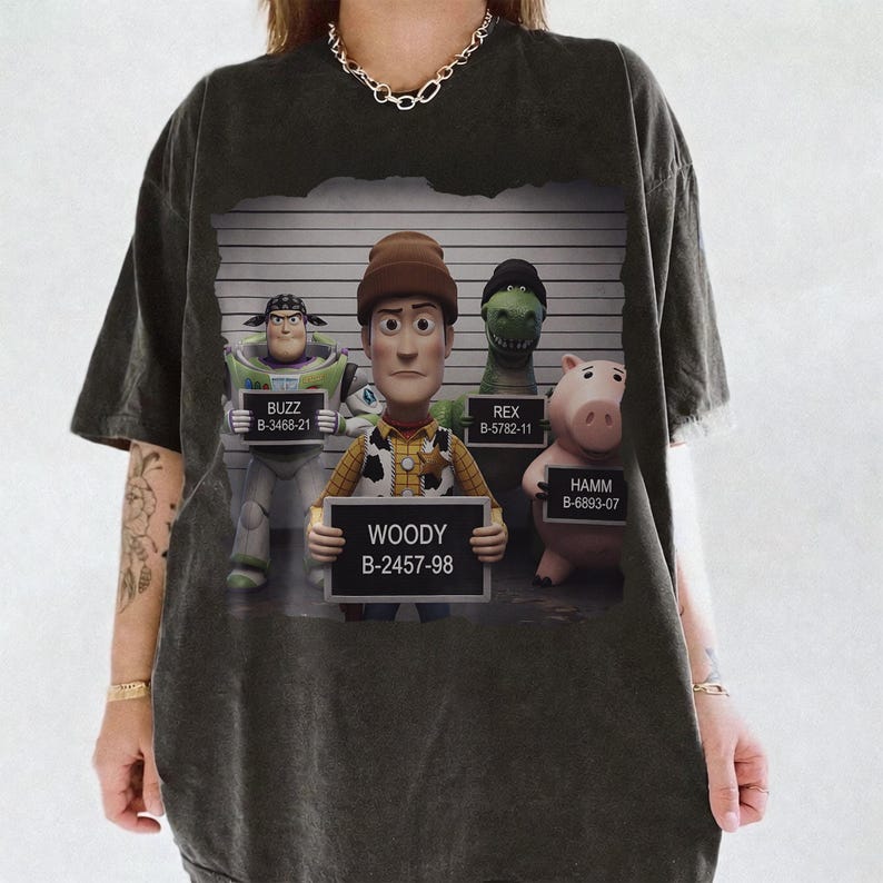 Puede incluir: Una camiseta gris carb&oacute;n con un estampado gr&aacute;fico de cuatro personajes de Toy Story posando para una foto policial frente a una tabla de altura. Los personajes Buzz, Woody, Rex y Hamm sostienen carteles con sus nombres y n&uacute;meros de identificaci&oacute;n.