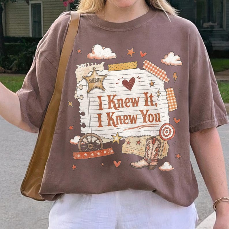 May include: A brown short-sleeve t-shirt featuring a graphic print of a torn piece of lined paper with the text "I Knew It, I Knew You" in brown letters, surrounded by illustrations of a sheriff star, a cowboy boot, a wagon wheel, and hearts.
