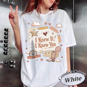May include: A white short-sleeve t-shirt featuring a graphic print of a lined notepad page with the text "I Knew It, I Knew You". The design includes a sheriff star, a cowboy boot, a wagon wheel, hearts, stars, and clouds in orange and brown.