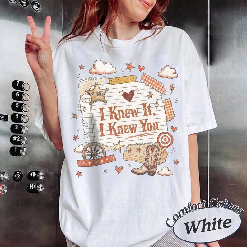 May include: A white short-sleeve t-shirt featuring a graphic print of a lined notepad page with the text "I Knew It, I Knew You". The design includes a sheriff star, a cowboy boot, a wagon wheel, hearts, stars, and clouds in orange and brown.