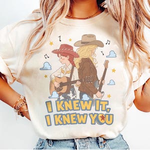May include: A cream-colored t-shirt featuring a graphic of two figures with guitars, one wearing a red cowboy hat and the other a brown hat. The text "I KNEW IT, I KNEW YOU" is printed in yellow with blue outlines below the figures, surrounded by stars and clouds.
