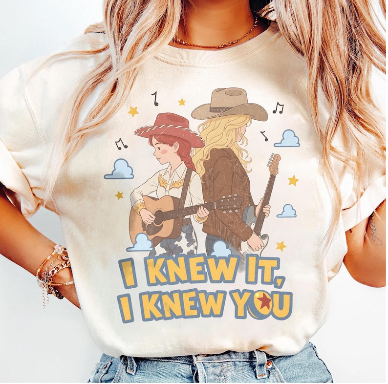 May include: A cream-colored t-shirt featuring a graphic of two figures with guitars, one wearing a red cowboy hat and the other a brown hat. The text "I KNEW IT, I KNEW YOU" is printed in yellow with blue outlines below the figures, surrounded by stars and clouds.