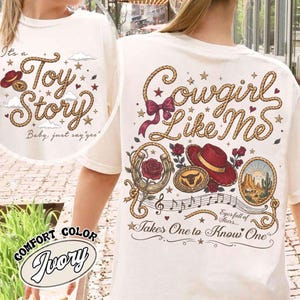 Pu&ograve; includere: Una t-shirt color avorio con un design grafico composto da scritte a forma di corda che recitano Cowgirl Like Me, accompagnate da illustrazioni di un cappello rosso, un ferro di cavallo con una rosa, una fibbia da toro, un deserto e stelle.