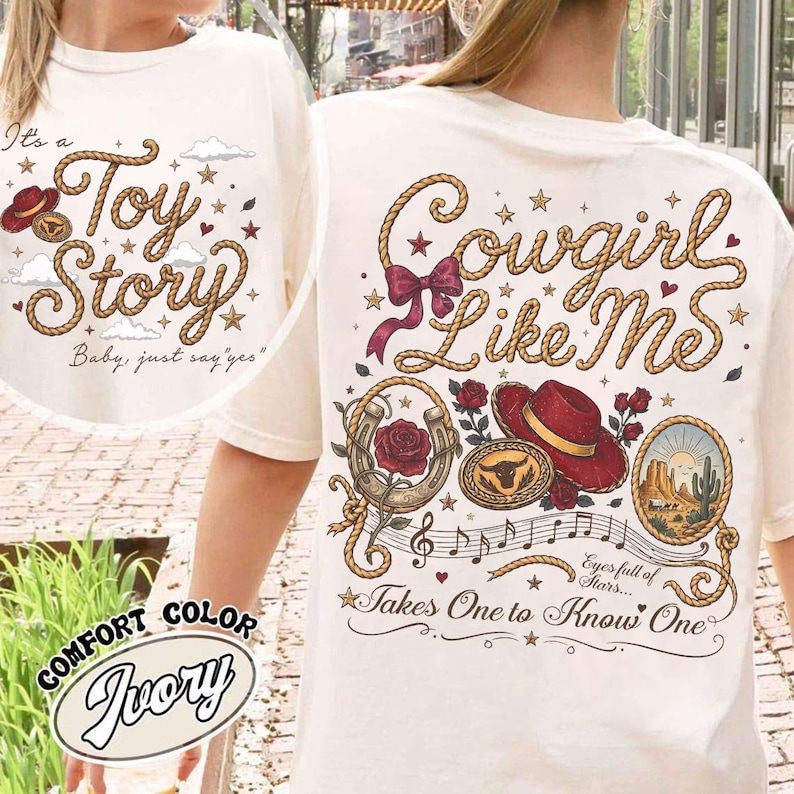 Pu&ograve; includere: Una t-shirt color avorio con un design grafico composto da scritte a forma di corda che recitano Cowgirl Like Me, accompagnate da illustrazioni di un cappello rosso, un ferro di cavallo con una rosa, una fibbia da toro, un deserto e stelle.