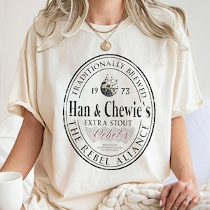May include: A cream-coloured t-shirt with a circular graphic. The graphic features the text "Han & Chewie's EXTRA STOUT Rebel's" in black and red, a ship illustration, and the words "Traditionally Brewed 1973".