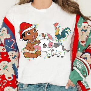Puede incluir: Camiseta blanca con una ilustración de dibujos animados de un bebé con gorro de Papá Noel, un cerdo con cuernos de reno y un gallo, todos adornados con luces navideñas. La camiseta se combina con un suéter estampado en rojo y verde.