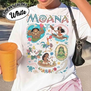 May include: White t-shirt with the word "MOANA" in colourful letters. The shirt features cartoon illustrations of characters from the movie, including Maui, Moana, Hei Hei, Pua, and others. The shirt also has the words "COMFORT COLORS" and "White" in a circle.