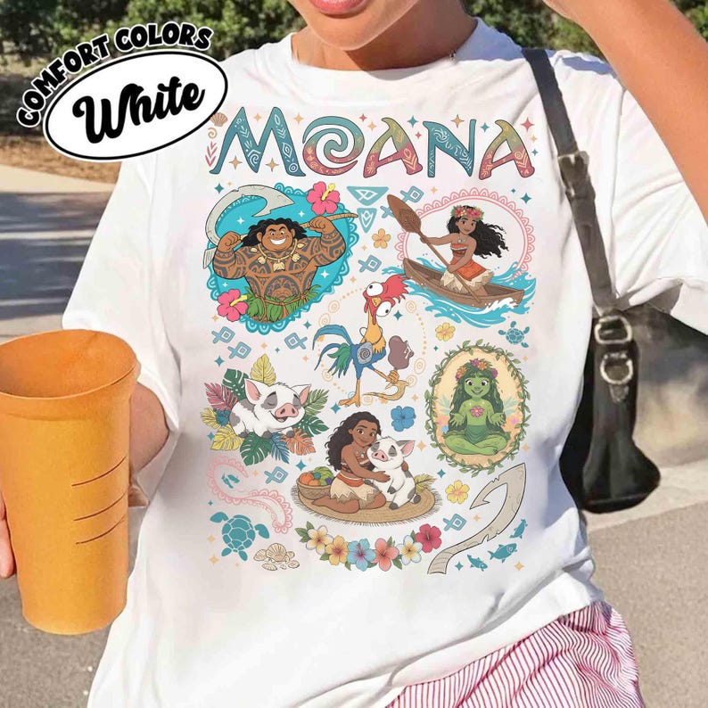 May include: White t-shirt with the word "MOANA" in colourful letters. The shirt features cartoon illustrations of characters from the movie, including Maui, Moana, Hei Hei, Pua, and others. The shirt also has the words "COMFORT COLORS" and "White" in a circle.