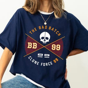 Puede incluir: Camiseta azul marino con un diseño gráfico que presenta una calavera blanca, las palabras "The Bad Batch", "BB 99" y "Clone Force 99". El diseño es en rojo, dorado y blanco. La camiseta tiene mangas cortas.