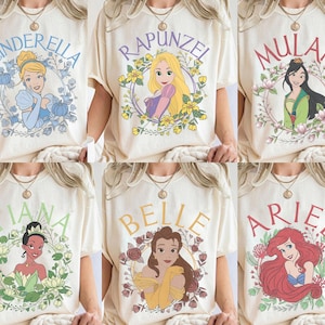 May include: A collection of six cream-colored t-shirts, each featuring a different illustrated princess character surrounded by a floral wreath and their name printed in an arched font: Cinderella, Rapunzel, Mulan, Tiana, Belle, and Ariel.