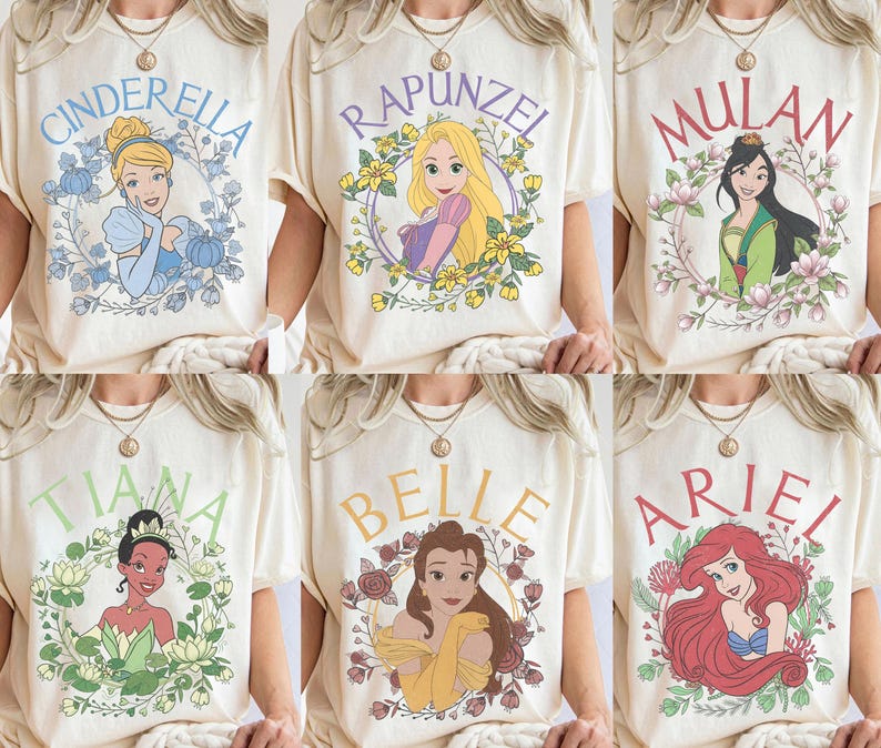 May include: A collection of six cream-colored t-shirts, each featuring a different illustrated princess character surrounded by a floral wreath and their name printed in an arched font: Cinderella, Rapunzel, Mulan, Tiana, Belle, and Ariel.