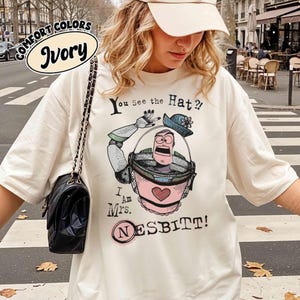 Puede incluir: Camiseta de color marfil con un dibujo animado de un personaje con un casco espacial rosa y gris, con el texto "You See the Hat?!" y "I Am Mrs. Nesbitt!" impreso en la parte delantera. La camiseta tambi&eacute;n tiene las palabras "Comfort Colors" e "Ivory" en un c&iacute;rculo.