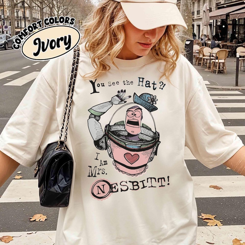 Puede incluir: Camiseta de color marfil con un dibujo animado de un personaje con un casco espacial rosa y gris, con el texto "You See the Hat?!" y "I Am Mrs. Nesbitt!" impreso en la parte delantera. La camiseta tambi&eacute;n tiene las palabras "Comfort Colors" e "Ivory" en un c&iacute;rculo.