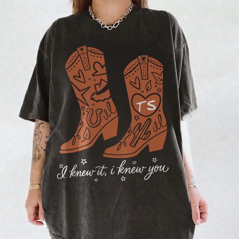 May include: A charcoal gray short-sleeve t-shirt featuring a graphic of two brown cowboy boots decorated with hearts, stars, and a horseshoe. One boot displays the letters TS inside a heart. The text "I knew it, I knew you" is printed below.