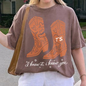 May include: A brown short-sleeve t-shirt featuring a graphic of two orange cowboy boots decorated with hearts, stars, and a horseshoe. The right boot displays the letters TS inside a heart. The text "I knew it, I knew you" is printed below.