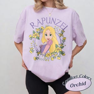 May include: A light purple short-sleeve t-shirt featuring a graphic of the character Rapunzel with long blonde hair, surrounded by a circular frame of yellow flowers and green leaves. The text RAPUNZEL is printed in a purple arc above the character.