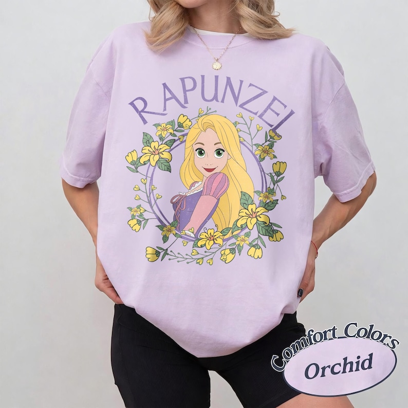 May include: A light purple short-sleeve t-shirt featuring a graphic of the character Rapunzel with long blonde hair, surrounded by a circular frame of yellow flowers and green leaves. The text RAPUNZEL is printed in a purple arc above the character.
