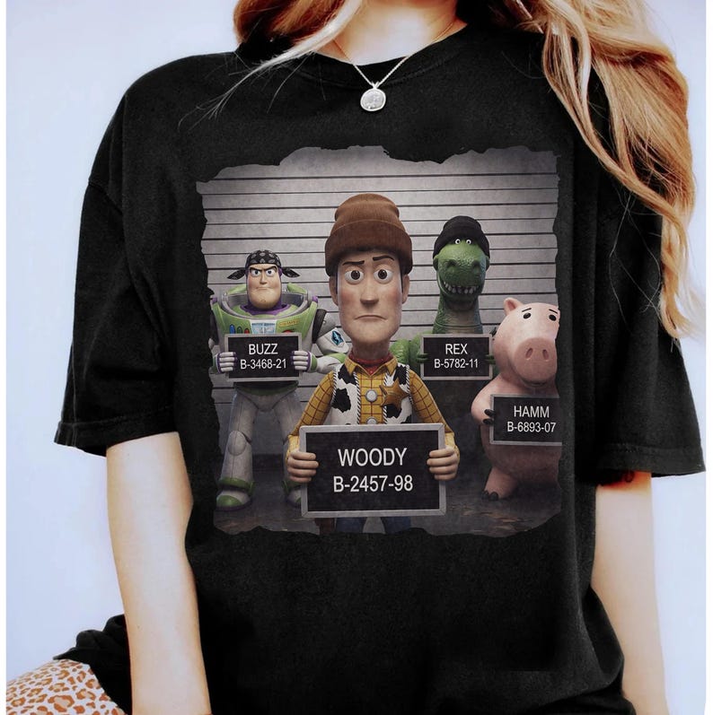 Puede incluir: Una camiseta negra con un estampado gr&aacute;fico de cuatro personajes de Toy Story posando para fotos policiales frente a una tabla de altura. Buzz, Woody, Rex y Hamm sostienen cada uno un cartel con su nombre y un n&uacute;mero de identificaci&oacute;n &uacute;nico.