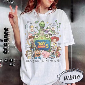 May include: A white short-sleeve t-shirt featuring a colorful graphic print of various animated characters from Toy Story. The design includes the text "TOY STORY 5" and "YOU'VE GOT A FRIEND IN ME" along with illustrations of Woody, Buzz Lightyear, Jessie, Rex, and other characters.